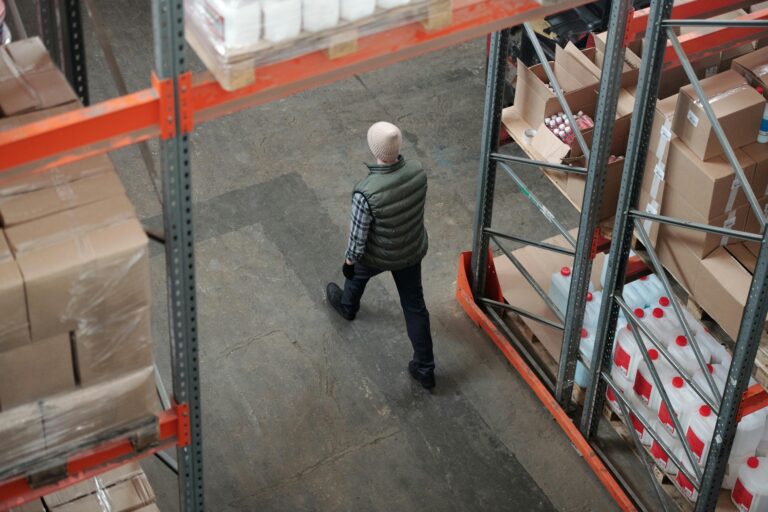 Overhead view of a worker walking through a warehouse aisle surrounded by shelves of packages.